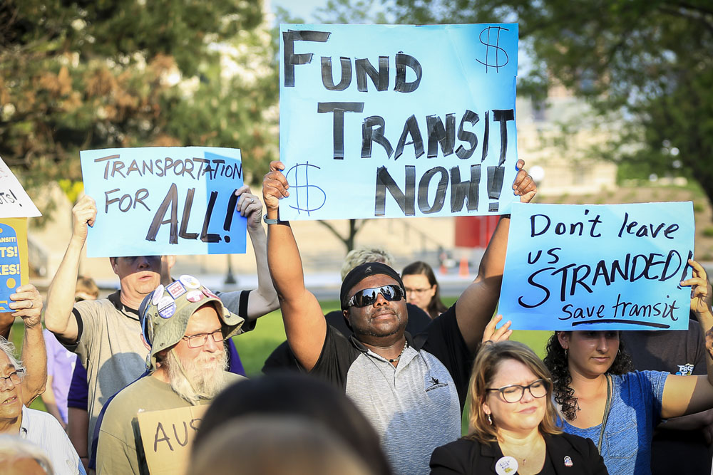 A group of people hold handmade signs at a public rally advocating for public transit funding. Signs read "FUND TRANSIT NOW!" "TRANSPORTATION FOR ALL!" and "Don’t leave us STRANDED! Save transit."